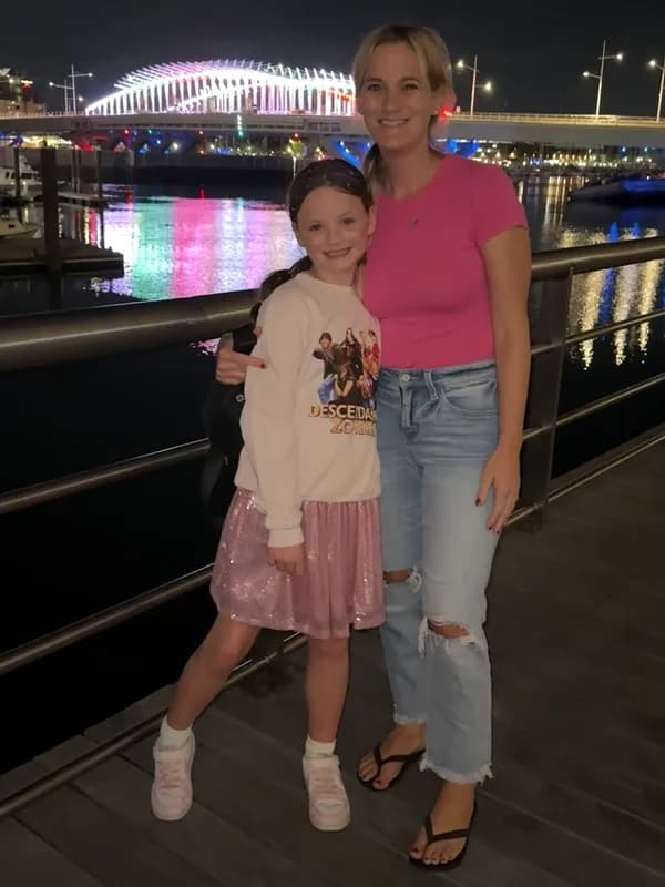 Woman and girl smiling by a waterfront at night with colorful bridge lights behind them.